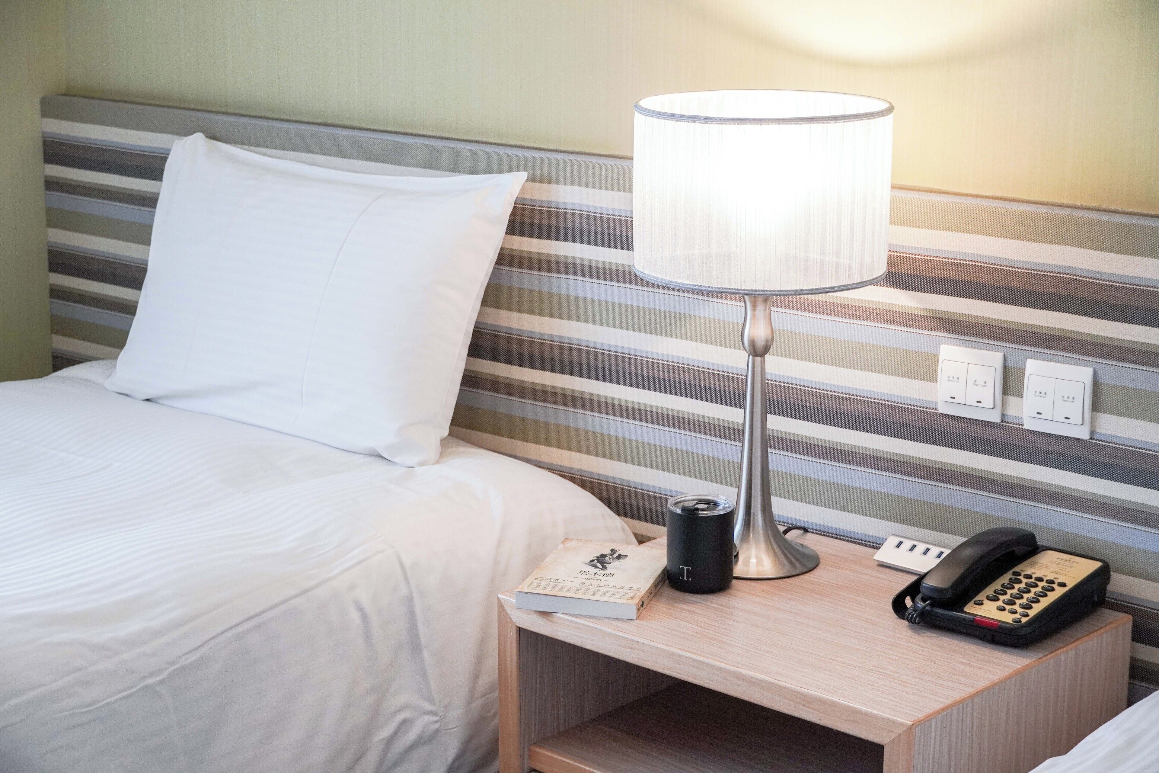 A hotel room with a bed, a bedside table with a lamp, a book, a phone, and a power outlet.