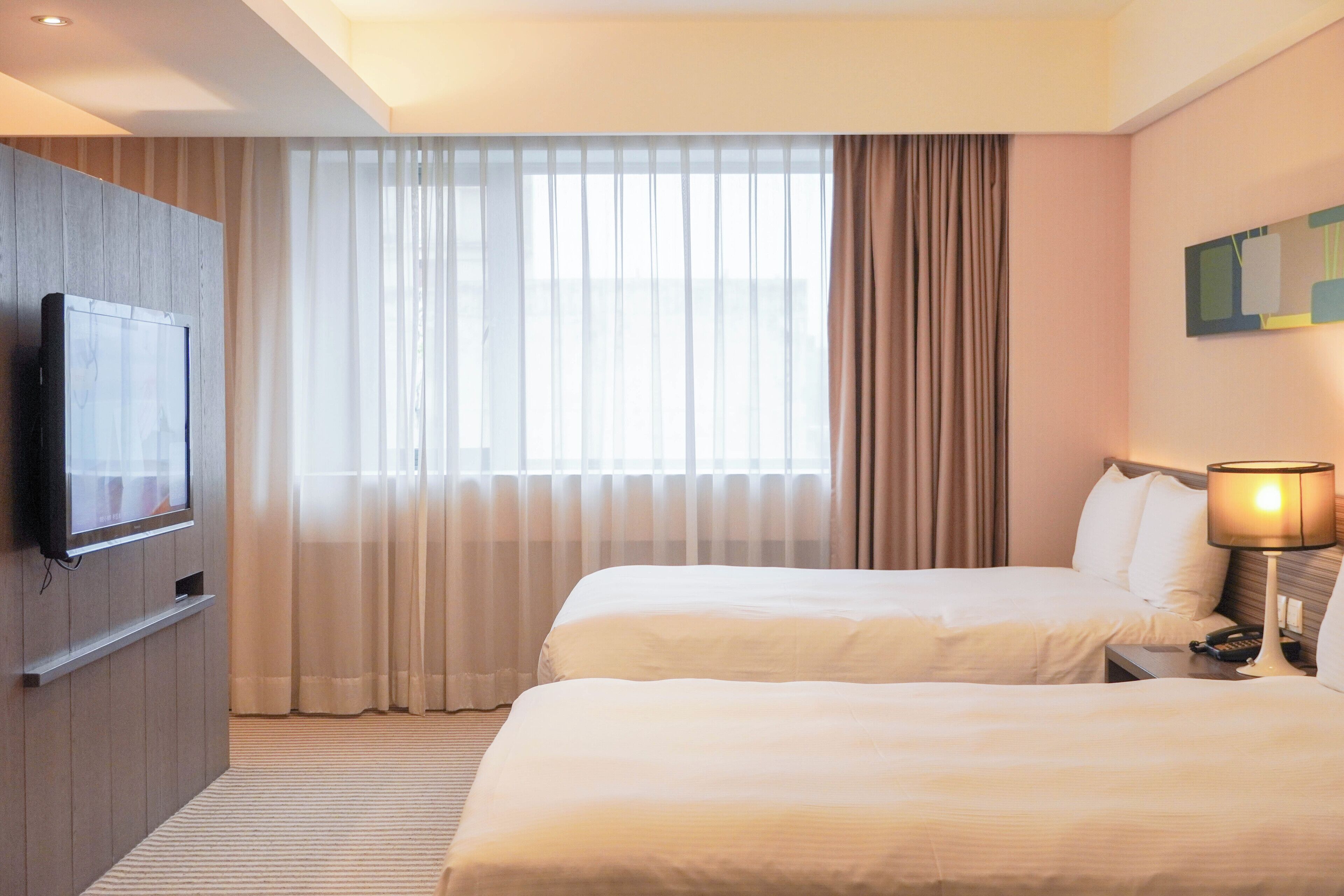 Une chambre d’hôtel avec deux lits, un téléviseur fixé au mur et une grande fenêtre avec des rideaux.