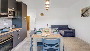 A modern kitchen with dark cabinets, a wooden countertop, and a built-in oven. There is a dining table set with plates, glasses, and a bottle. A gray sofa is in the background, and a wall clock is visible. - La Dolce Laveno, Balcony Apartment With A/C (Laveno Mombello)