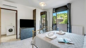 A modern bedroom with a bed, a television, a washing machine, and a view of a green landscape. - La Dolce Laveno, Balcony Apartment With A/C (Laveno Mombello)