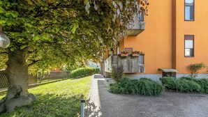 A paved pathway leading to a building with a yellow facade, surrounded by greenery and a tree with dense foliage. - La Dolce Laveno, Balcony Apartment With A/C (Laveno Mombello)