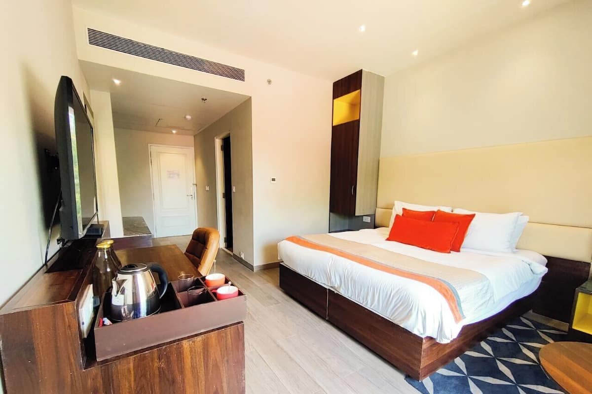 A hotel room with a bed, a desk with a kettle, a chair, and a TV.