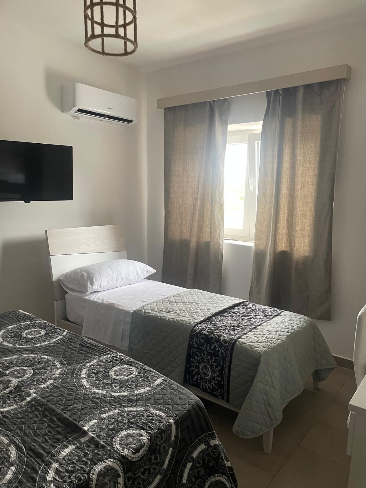 Ein Hotelzimmer mit zwei Einzelbetten, einem an der Wand befestigten Flachbildfernseher und einem Fenster mit Vorhängen.