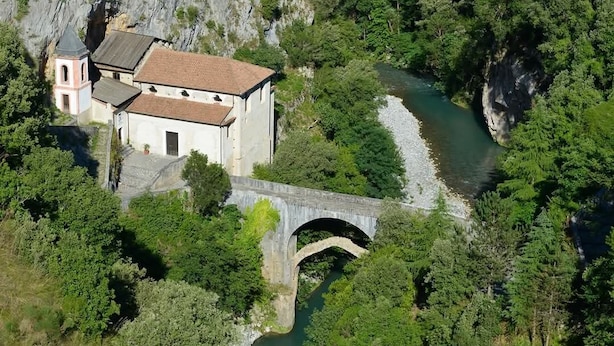 Eine Steinkonstruktion überspannt einen Fluss, umgeben von dichtem Grün und einem Gebäude mit einem Glockenturm.