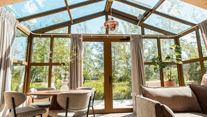 Un salón de cristal con techo de madera, amplios ventanales, una mesa de comedor con sillas y un sofá.