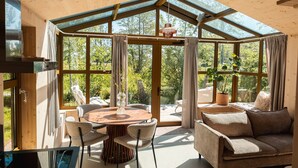 Una habitación acogedora con techo de madera, una mesa redonda de madera con sillas, un sofá y ventanas amplias con vistas a árboles.