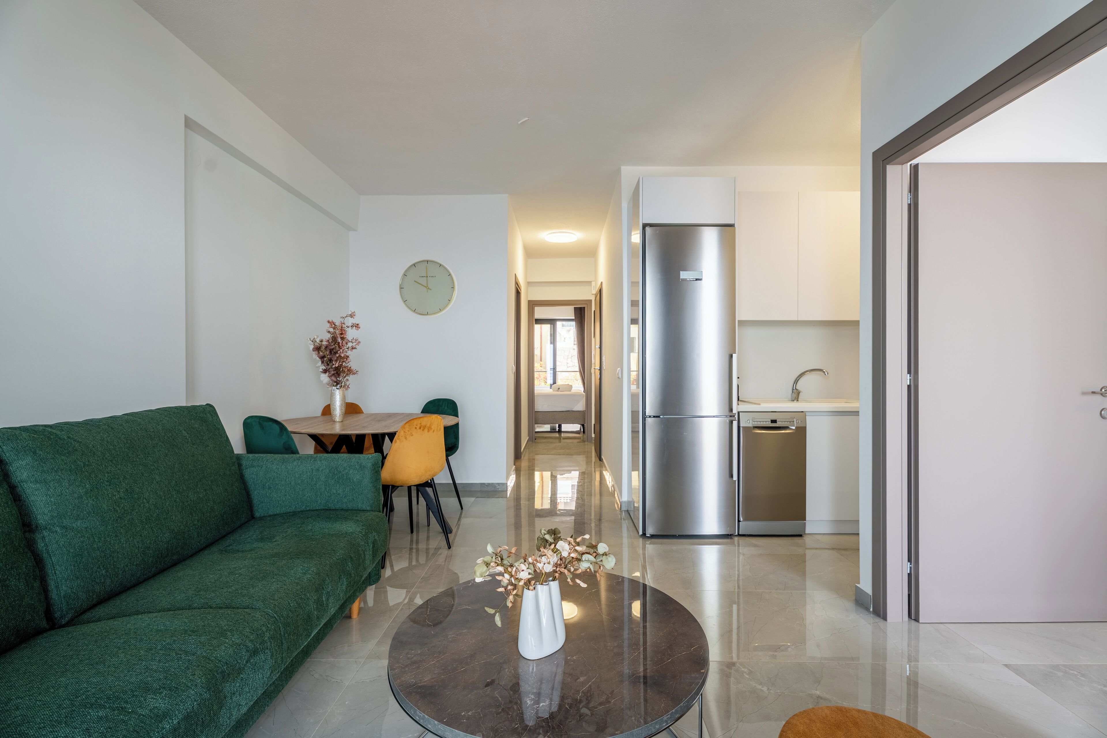 Un salon moderne avec un canapé vert, une table basse ronde surmontée d’un vase, et un coin repas avec des chaises et une table. On aperçoit au fond une cuisine équipée d’un réfrigérateur et d’un lave-vaisselle.