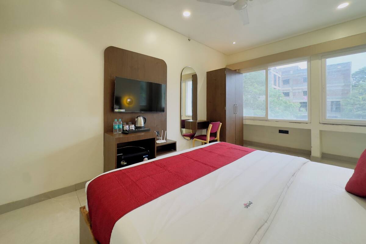 A hotel room with a large bed, a TV mounted on the wall, a wardrobe, a desk with a chair, and a window with a view of buildings.