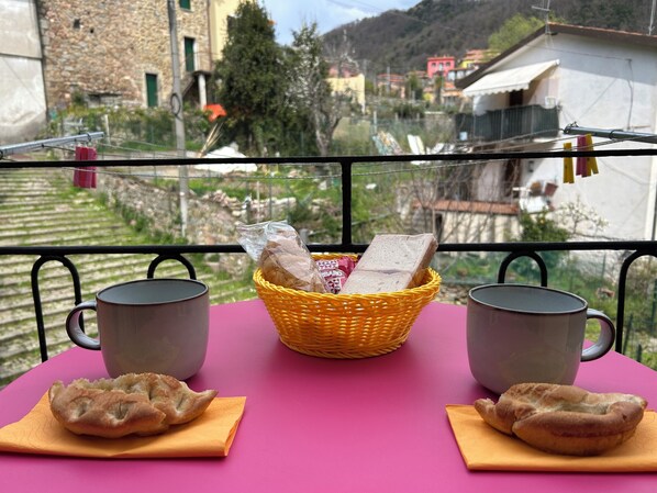 A balcony with a wicker basket containing bread, two mugs, and two pastries on a pink table.