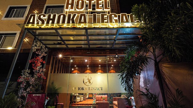 L’extérieur de l’hôtel Ashoka Legacy se distingue par une enseigne lumineuse bien visible, une entrée en verre et un espace de réception agrémenté de sièges.
