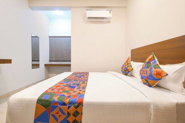 A hotel room with a bed, pillows, a blanket, an air conditioning unit, a window, and a mirror.