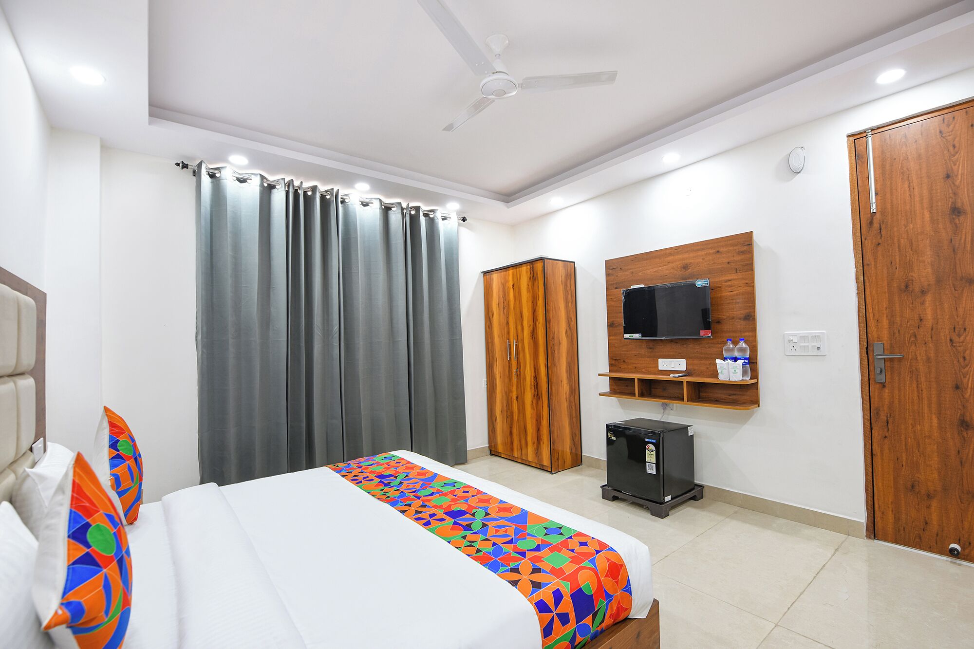 Habitación de hotel con cama, cortinas, ventilador de techo, un televisor montado en un panel de madera, un mueble pequeño y una puerta.