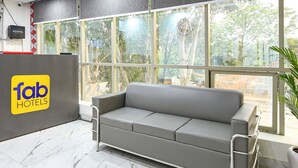 A hotel reception area with a grey sofa, a reception desk with a 'fab hotels' sign, and large windows offering a view of trees and buildings. - Fabhotel Evergreen (Faridabad)