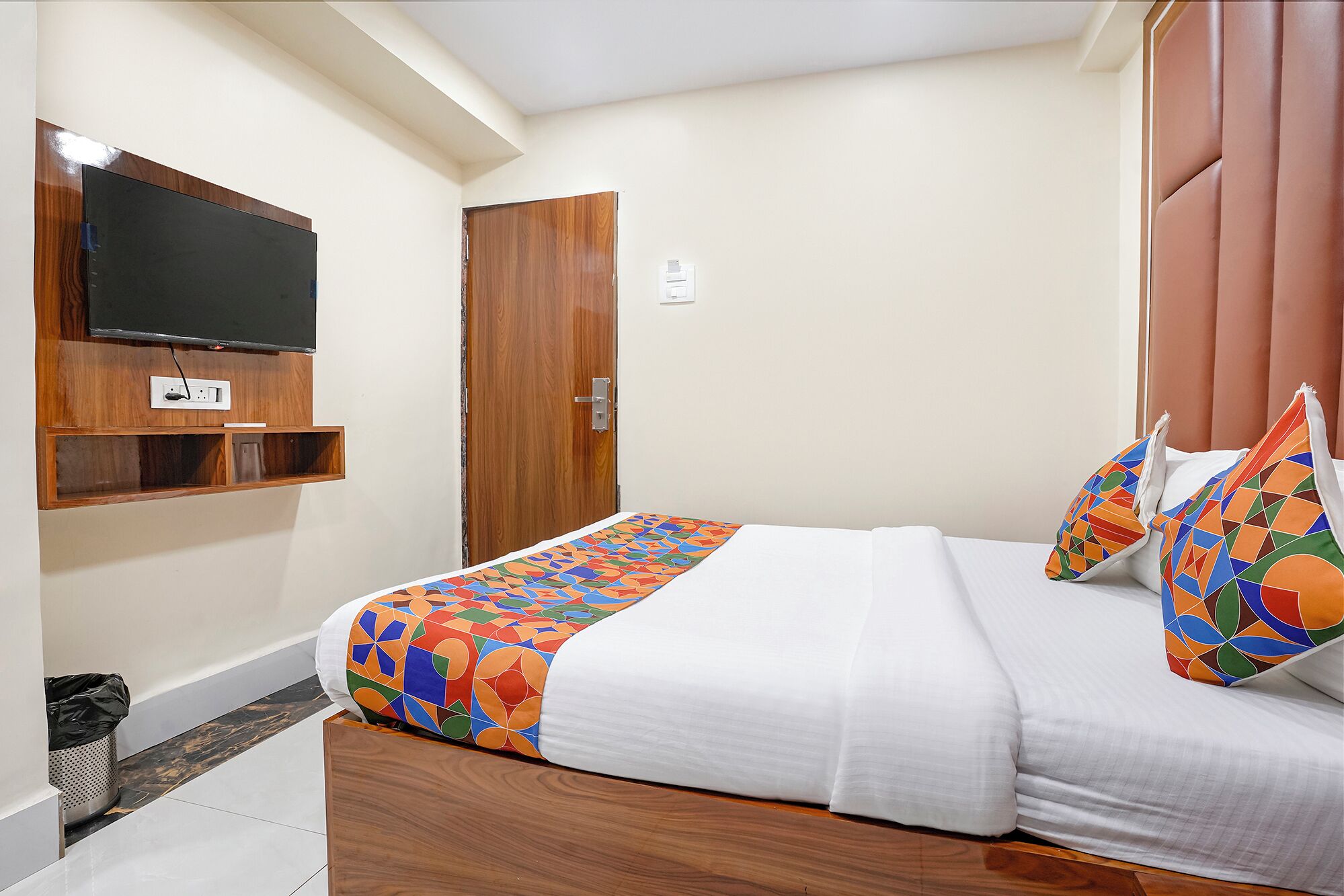 A hotel room with a bed, a television mounted on the wall, a wooden door, and a small shelf.