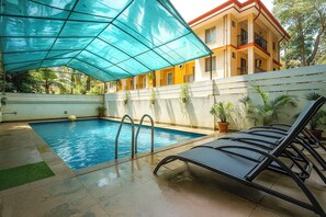 A swimming pool with a blue canopy, a lounge chair, and a building with balconies in the background. - Fabhotel Bagatel (Calangute)