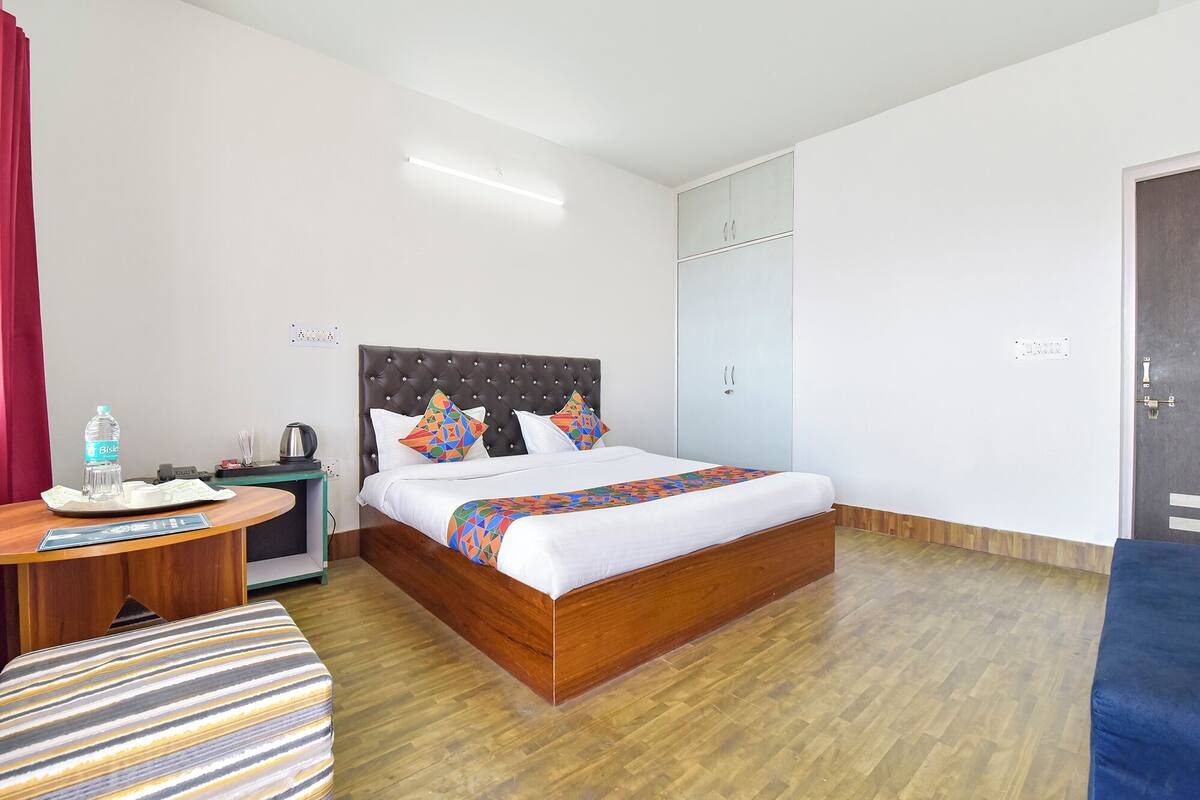 Habitació d'hotel amb un llit gran, una tauleta de nit de fusta, un otomà amb ratlles, una ampolla d'aigua i un escriptori amb una cadira.