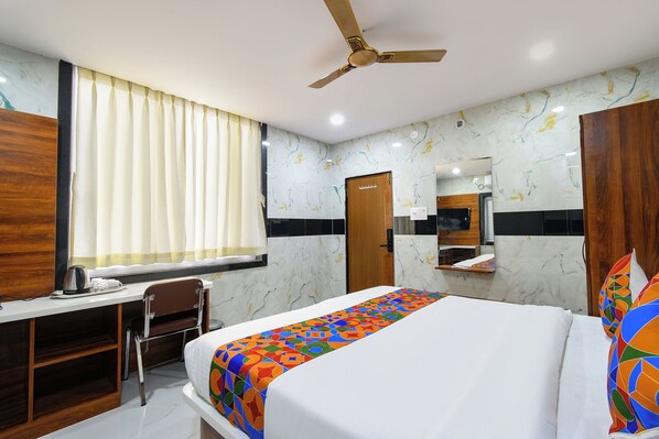 A hotel room with a bed, a desk, a chair, a ceiling fan, a window with curtains, and a bathroom with a mirror and sink. - Fabhotel Sunrise (Hyderabad)