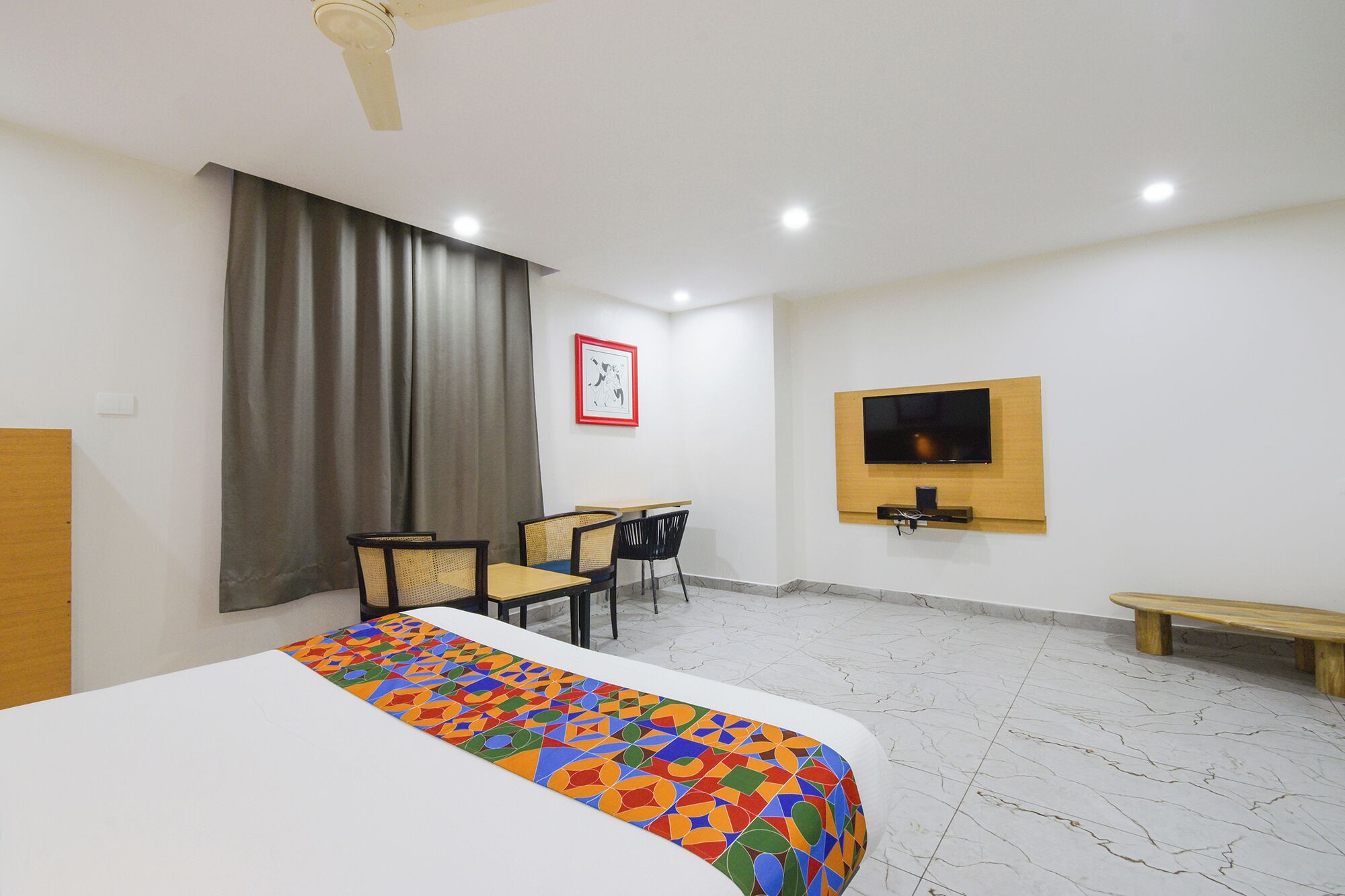 A hotel room with a bed, a desk, two chairs, a TV mounted on the wall, and a window with curtains.
