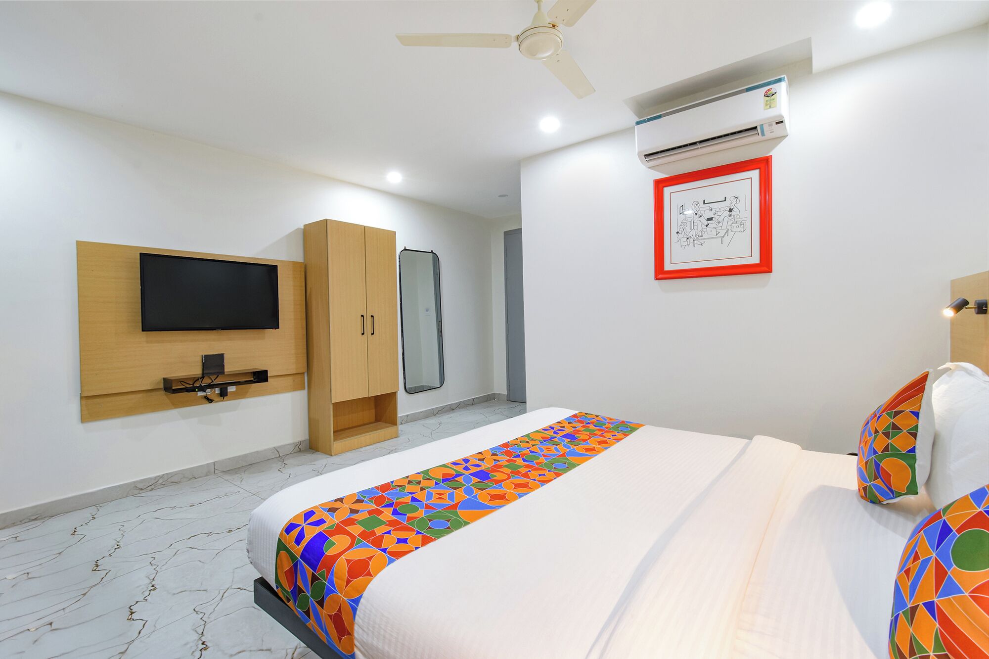 A hotel room with a bed, a TV mounted on the wall, a wardrobe, an air conditioning unit, and a framed picture.