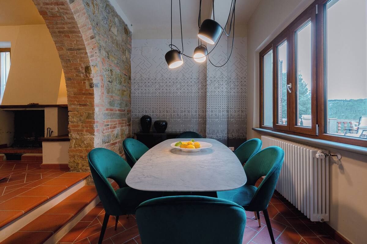 Uma sala de jantar com uma mesa de mármore, cadeiras verdes, uma cesta de frutas e vista através de amplas janelas.