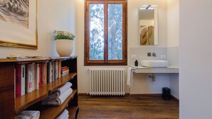 A bathroom with a wooden floor, a radiator, a sink, a mirror, and a window with a view of trees. - Casa Riccimballa (Impruneta)