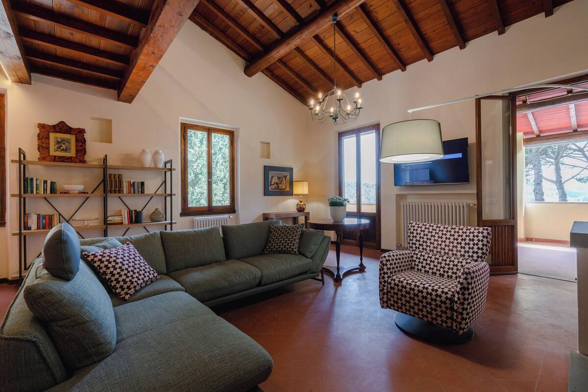 Uma sala de estar aconchegante com sofá seccional, poltrona estampada, vigas de madeira no teto, estantes e televisão.