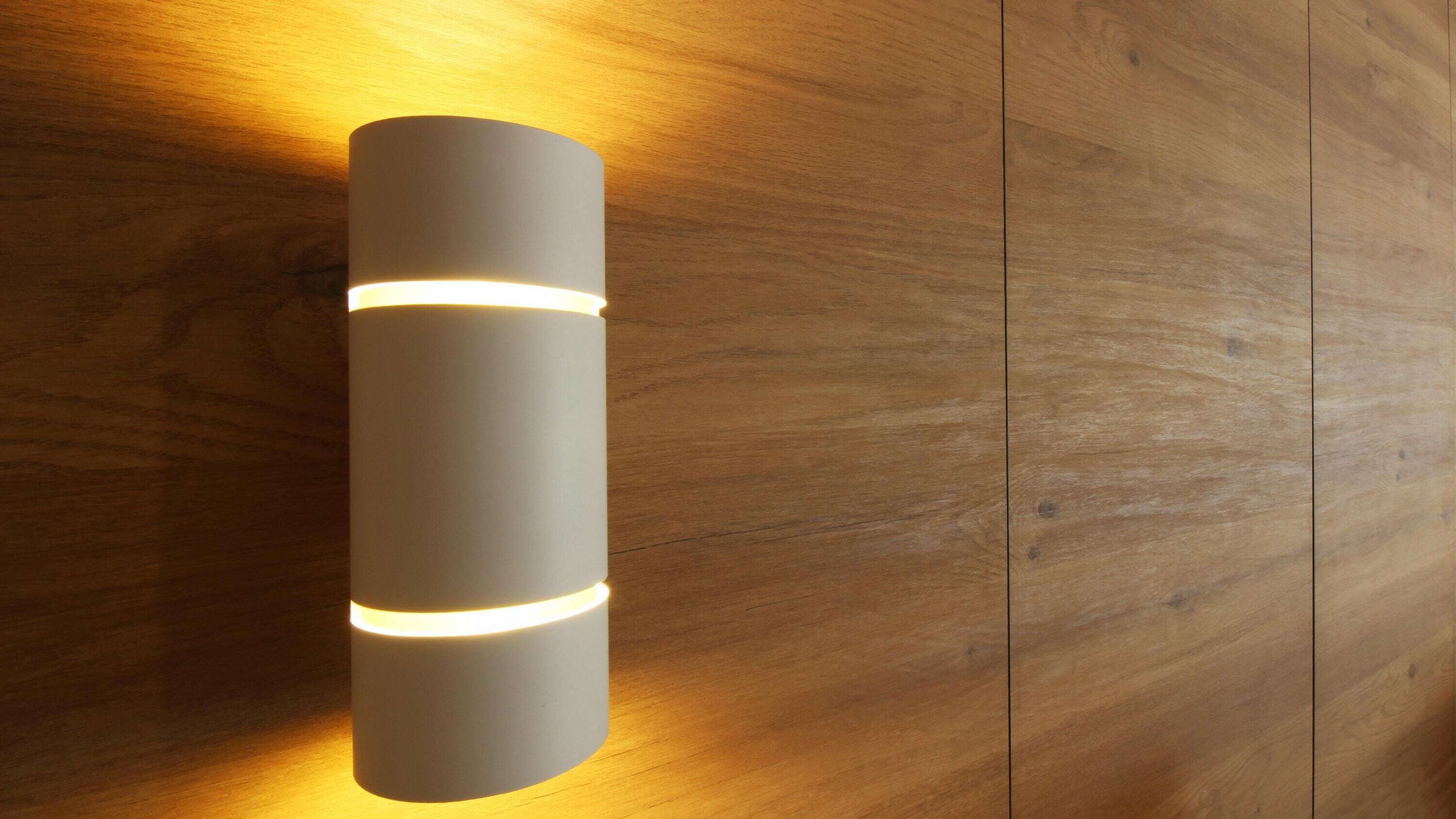 A cylindrical wall-mounted light fixture with two illuminated bands, set against a wooden panel background.