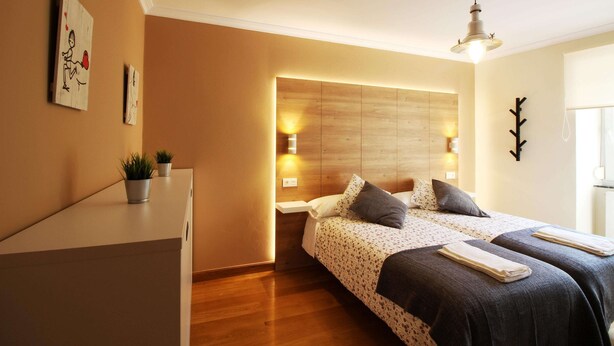 Une chambre d’hôtel moderne avec une tête de lit en bois, deux lits avec une literie à motifs, des lampes de chevet et un petit bureau agrémenté de plantes en pot.