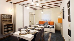 Un coin repas avec une table dressée pour quatre personnes, un buffet en bois avec de la verrerie, et un lit avec un oreiller orange.