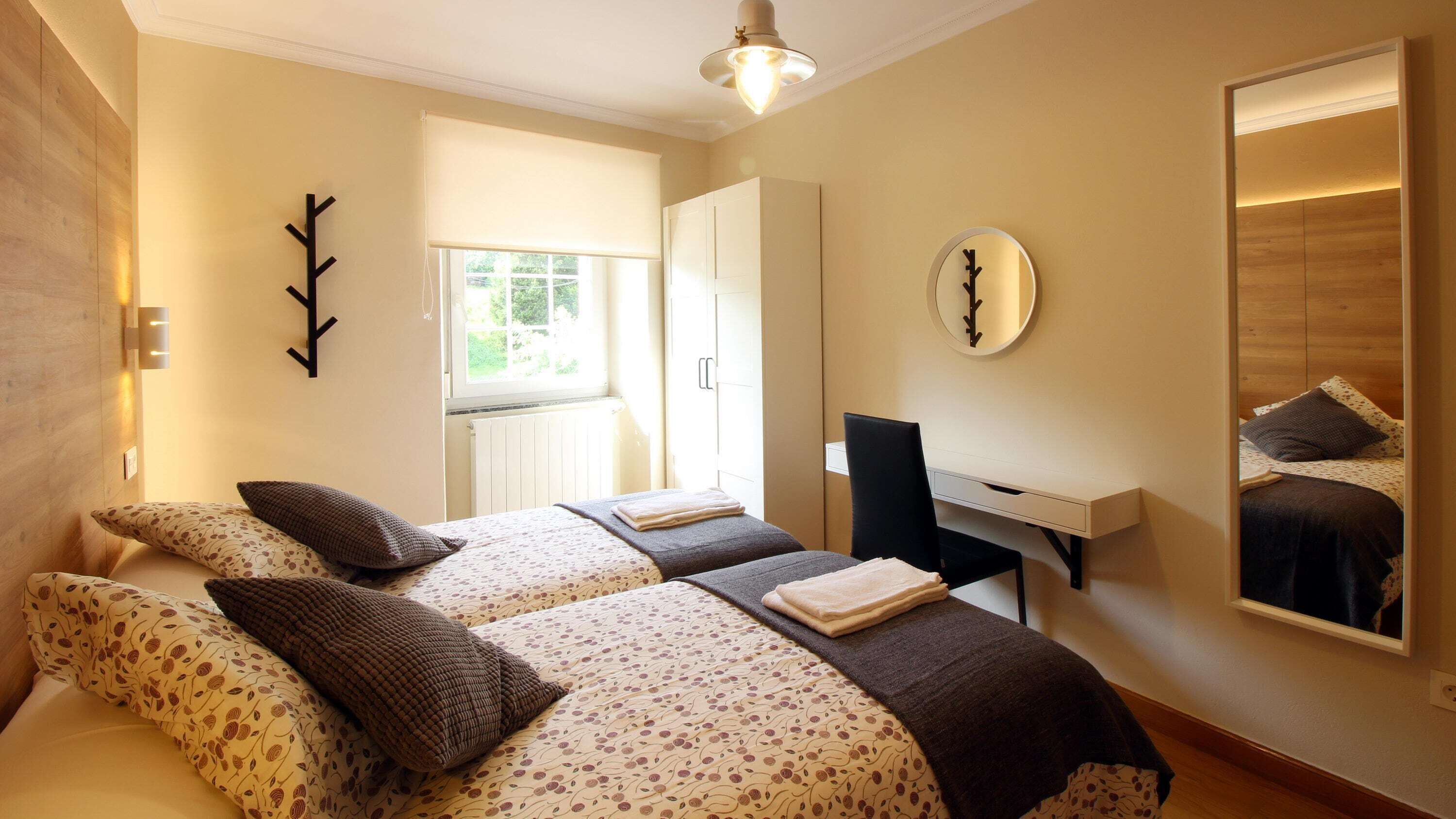 A bedroom with two beds, a desk, a mirror, and a window.