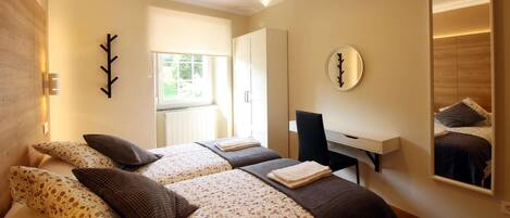 A bedroom with two beds, a desk, a mirror, and a window.