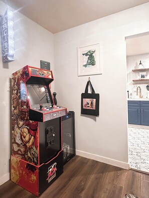 Una habitación con una máquina de juegos arcade, un cuadro con la imagen de un personaje, un bolso colgado en la pared y una zona de cocina con estantes.