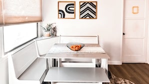 Un área de comedor con una mesa blanca, un banco y una alfombra. Hay dos obras de arte enmarcadas en la pared y una puerta de diseño sencillo.
