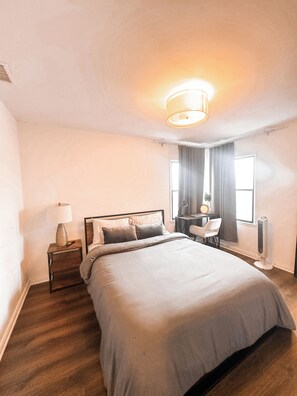 Une chambre d’hôtel avec un grand lit, une table de chevet, une lampe, un bureau, une chaise et un ventilateur sur pied.