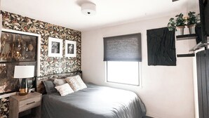 Un dormitorio con cama, mesita de noche, lámpara, ventana con persiana y un cuadro en la pared.