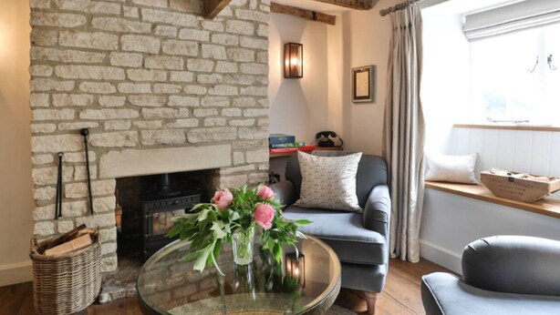 A cozy living room with a stone fireplace, a sofa, a coffee table with flowers, and a window with curtains.