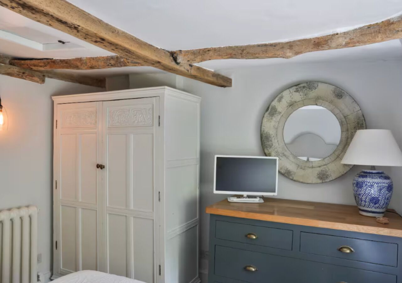 A room with a white wardrobe, a wooden dresser, a round mirror, a small TV, and a radiator.