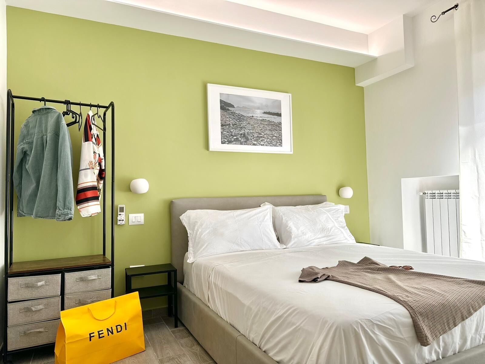 Una camera d'hotel con un letto, un comodino, un armadio e un appendiabiti con vestiti e una borsa della spesa.