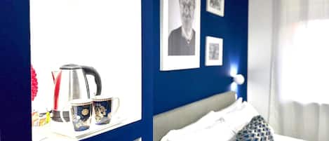 A hotel room with a bed, a nightstand with a kettle and mugs, a large mirror, and framed pictures on the wall.