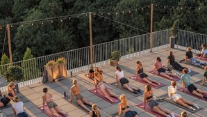 A group of people on yoga mats outdoors, arranged in rows on a paved surface with a railing and trees in the background.