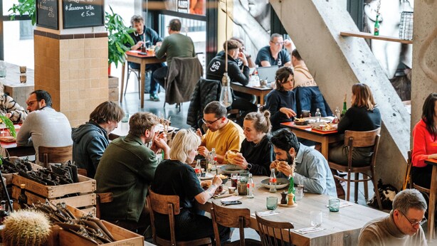 A bustling café with patrons seated at tables, surrounded by a modern interior with exposed concrete beams and large windows.