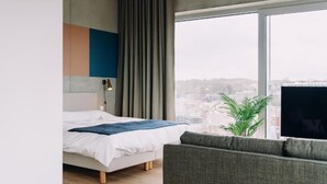 A modern hotel room with a bed, a grey sofa, a large window with a city view, and a plant.