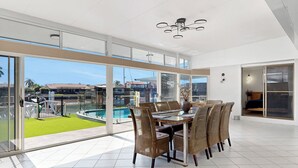 A spacious room with a large dining table and wicker chairs, a modern chandelier, and a view of a swimming pool and buildings. - Relfections at Newport 5Bedroom Pool and Canal Living (Newport)