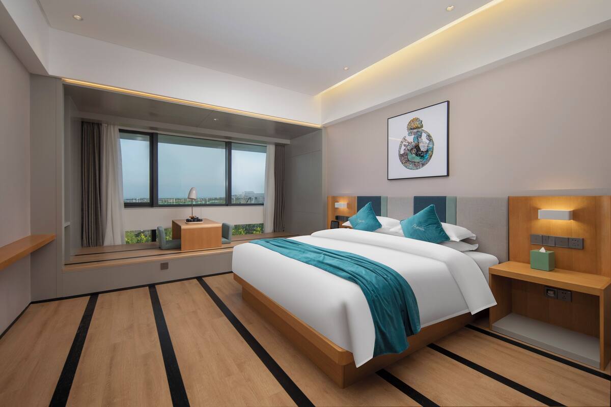 A modern hotel room with a large bed, a window with curtains, a bedside table, and a wall-mounted artwork.