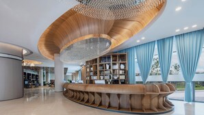 Un hall d’entrée moderne avec un comptoir d’accueil en bois aux lignes courbes, un lustre et de grandes fenêtres ornées de rideaux bleus.