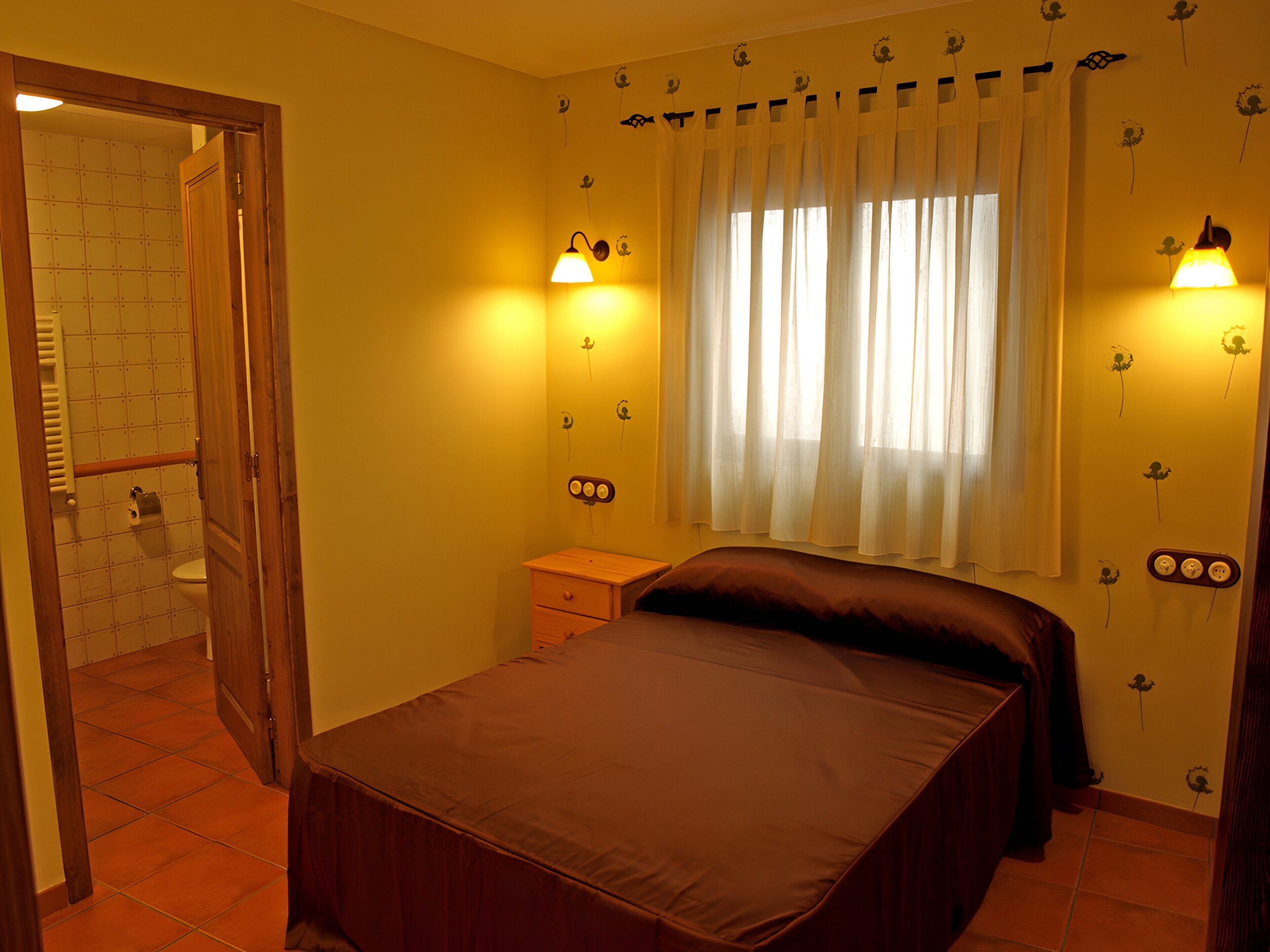 Un dormitorio con cama, mesita de noche, ventana con cortinas e iluminación en la pared.
