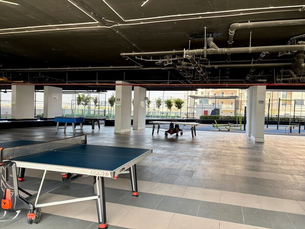 An indoor recreational area with ping pong tables, exercise equipment, and a view of the outside.