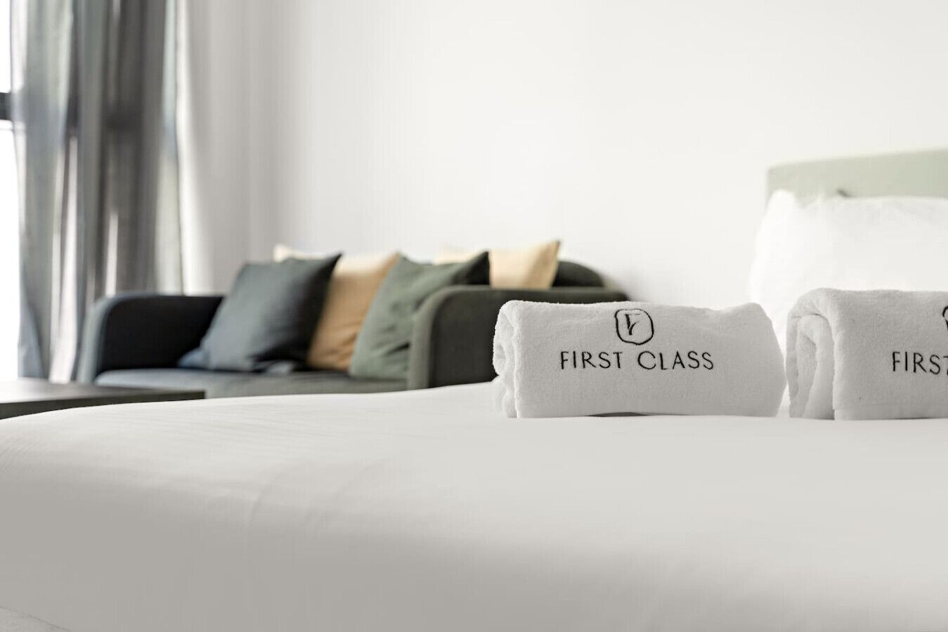 A neatly made bed with two folded towels labeled "FIRST CLASS" and a dark-colored sofa with cushions in a modern room.
