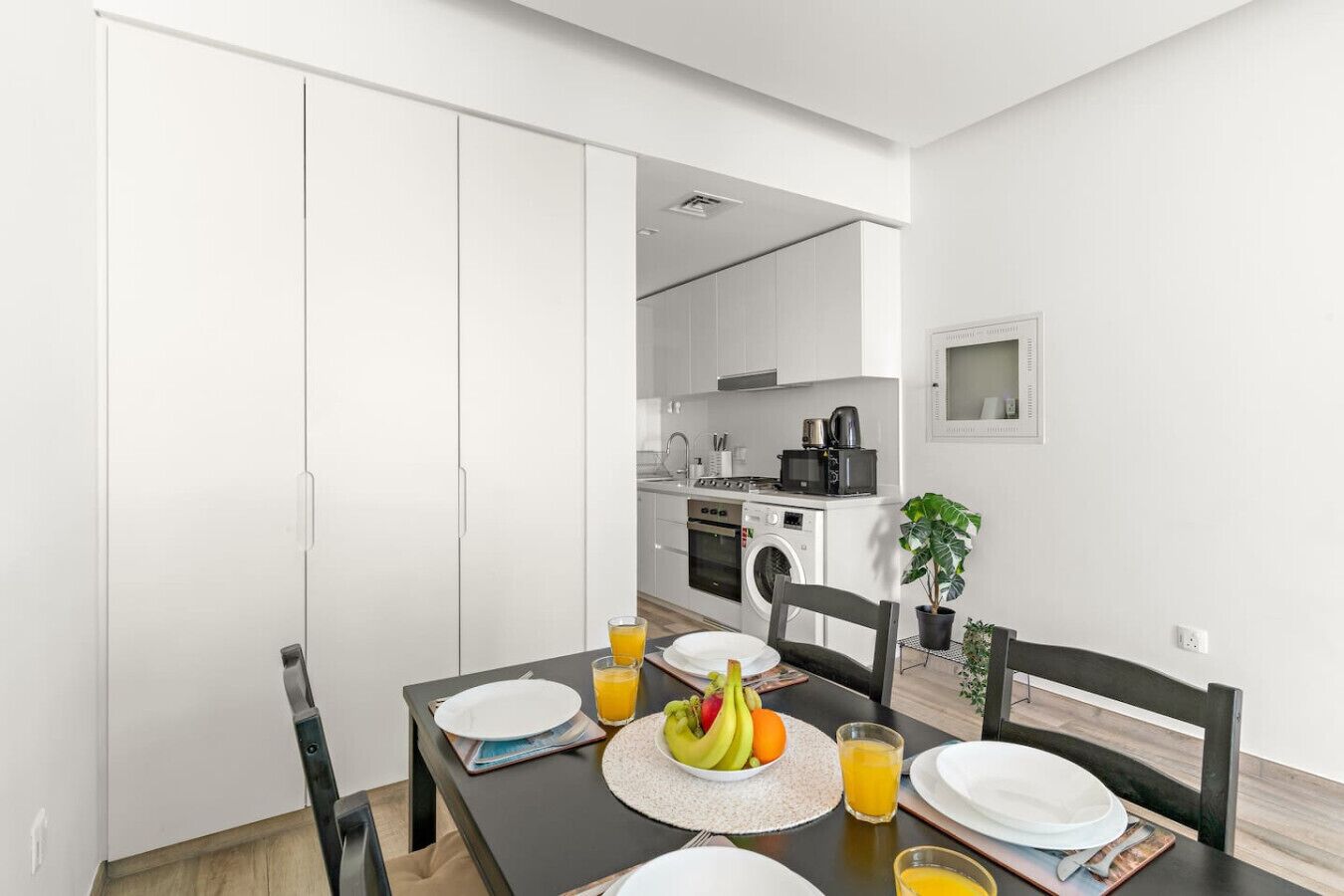 A modern kitchen with white cabinets, a microwave, and a coffee maker. There is a dining table set with plates, glasses, and fruit.
