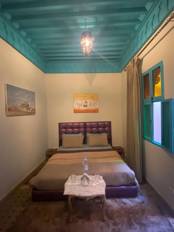 A bedroom with a bed, a table with glasses and a bottle, a painting, and a window with curtains. - Riad Massin Marrakech (Marrakech)